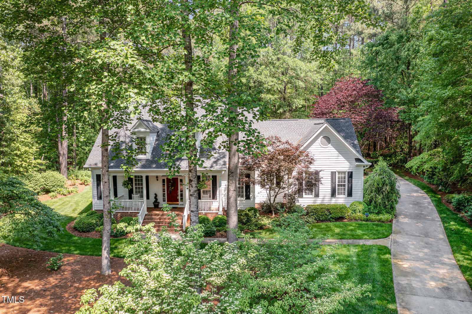 A white, single-story house with black shutters and a covered front porch sits surrounded by tall trees and lush greenery. A long driveway curves to the right of the house through the landscaped yard.