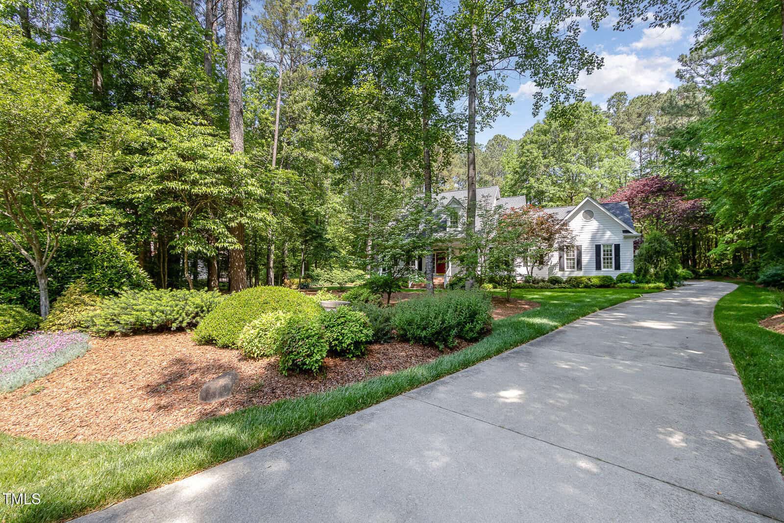 A long, curved driveway leads through lush, landscaped greenery and trees to a white house with a gabled roof, set in a wooded area under a blue sky.