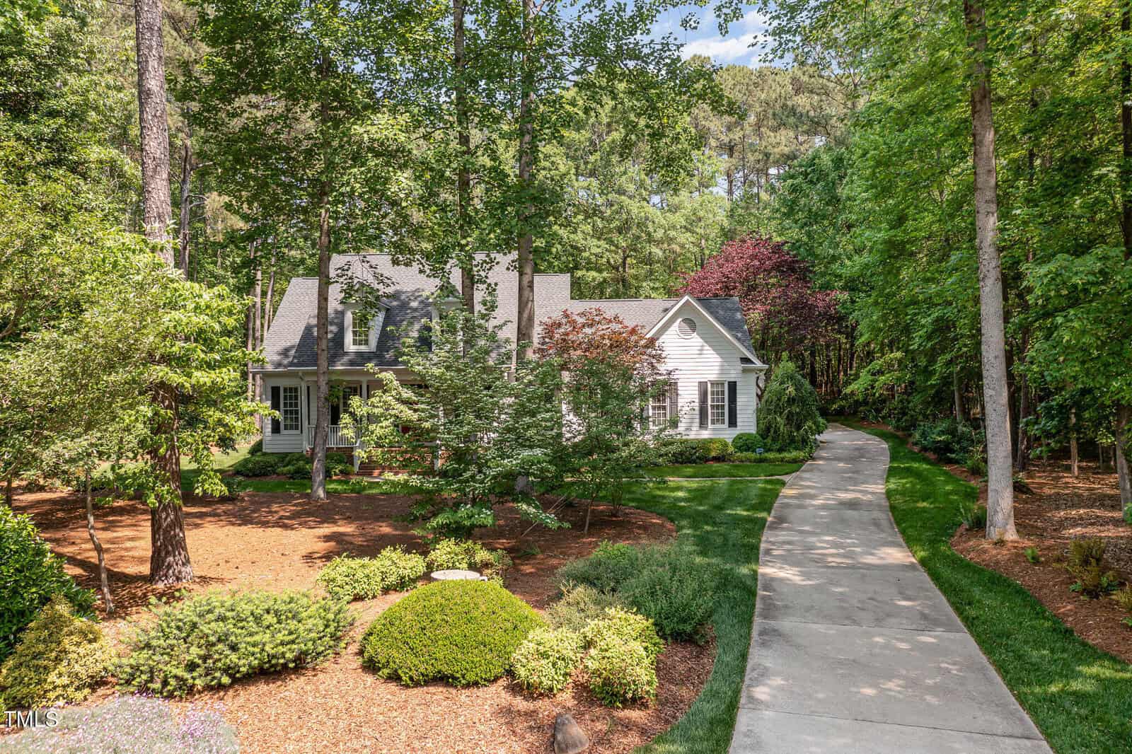 A white house with a covered porch is surrounded by trees and landscaped greenery. A curved concrete driveway leads from the street to the house, passing through well-maintained gardens and mulch beds.