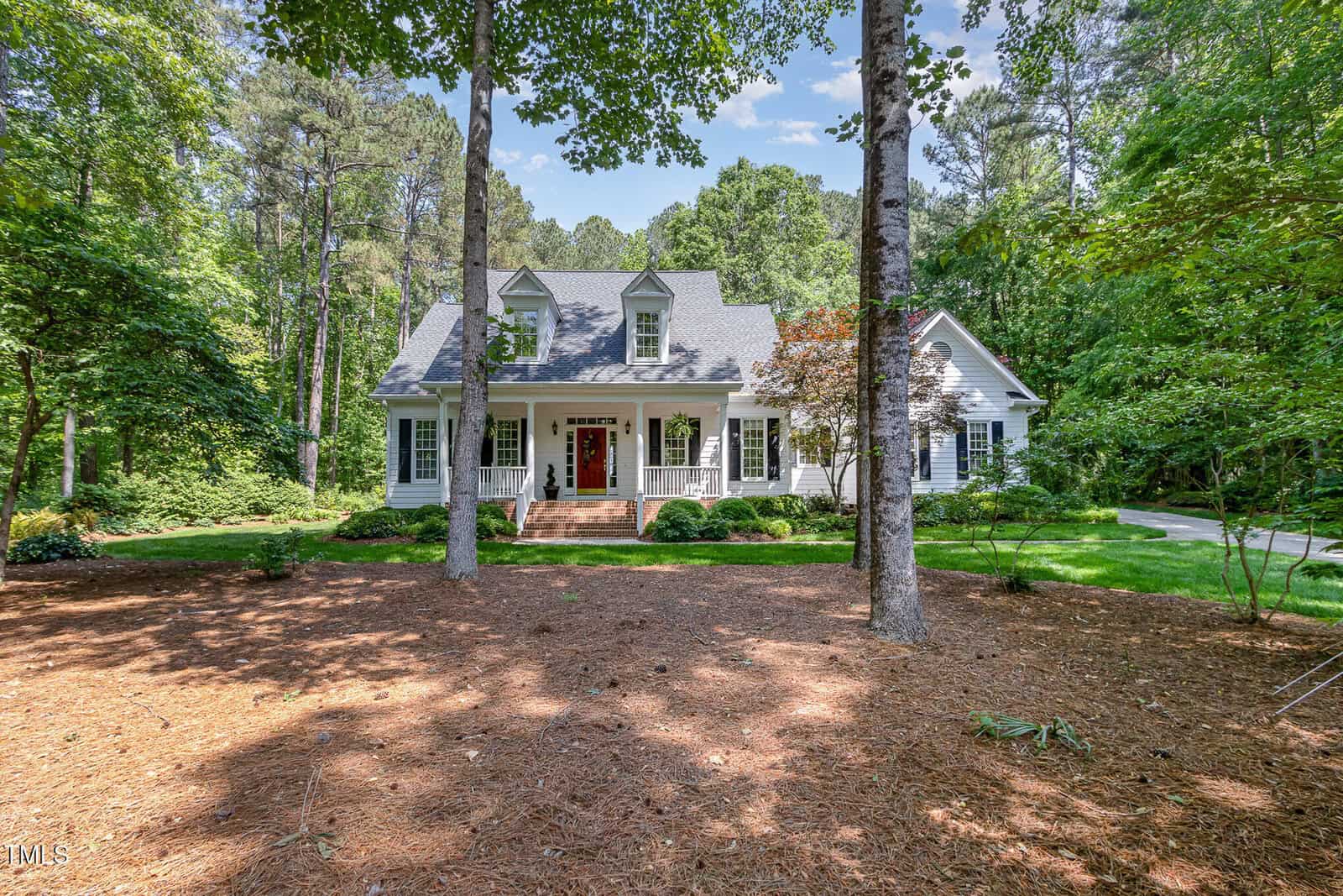 A white two-story house with a covered front porch, brick steps, black shutters, and dormer windows, surrounded by tall trees and greenery, set in a wooded area with a pine straw-covered yard.