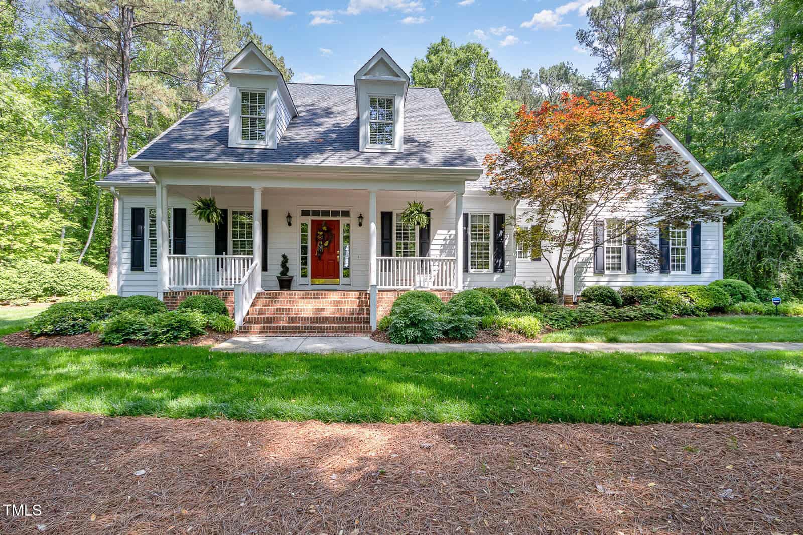 A white two-story house with dormer windows, a large front porch with columns, hanging ferns, red front door, brick steps, and a landscaped yard surrounded by trees.