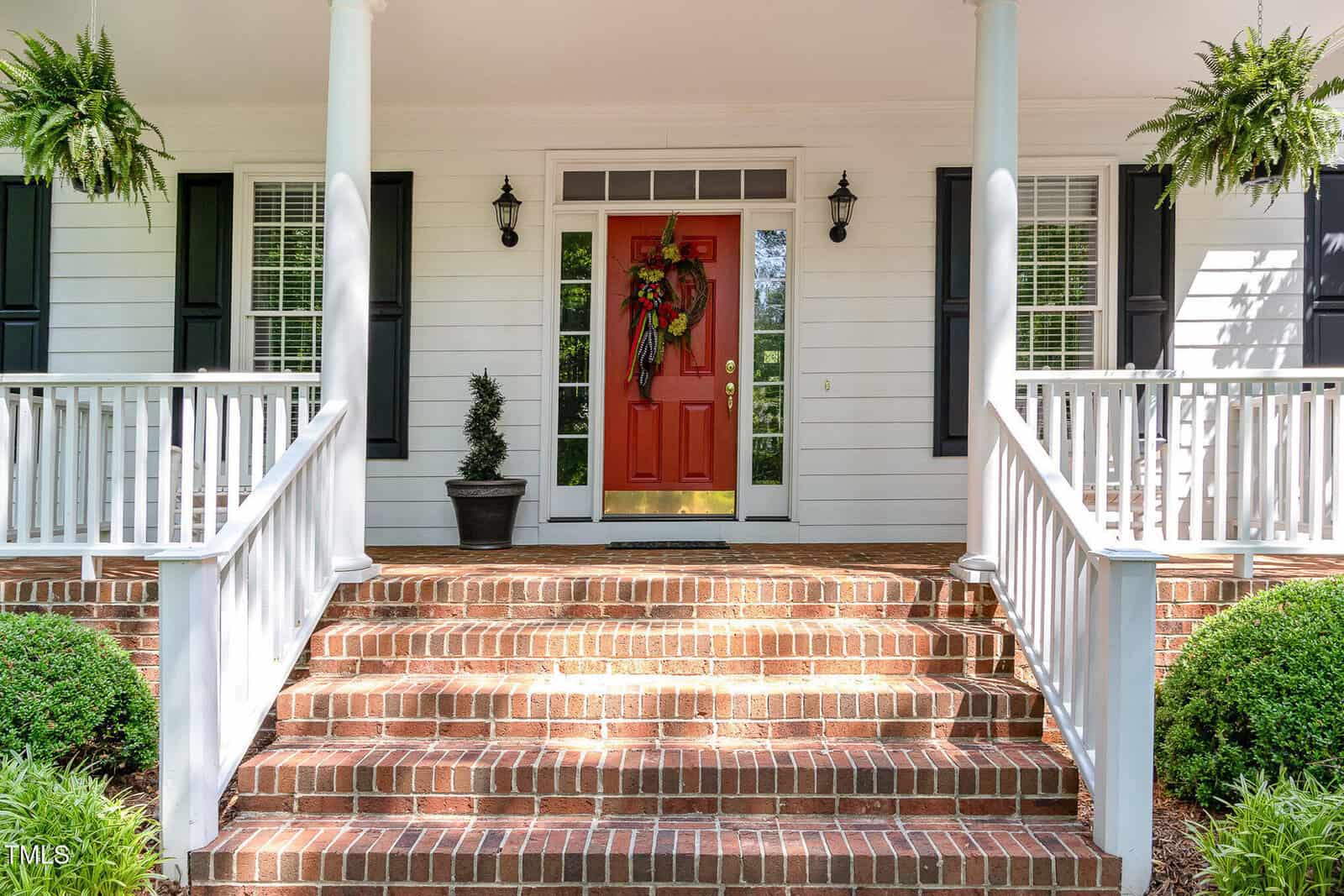 Brick steps lead to a porch with white railings and columns. The front door is red and features a decorative wreath. There are two potted plants and ferns hanging from the porch ceiling. White siding and windows with shutters are visible.