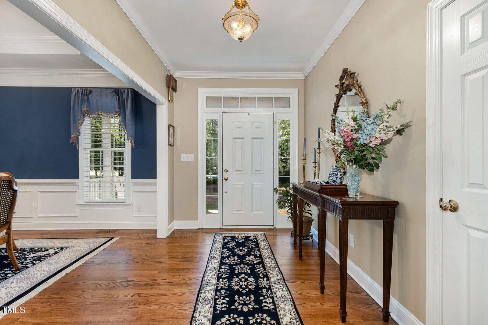 A hallway with hardwood floors, a white front door with windows, a console table with a mirror and floral arrangement, and a blue patterned runner rug. A dining room with blue walls is visible to the left.