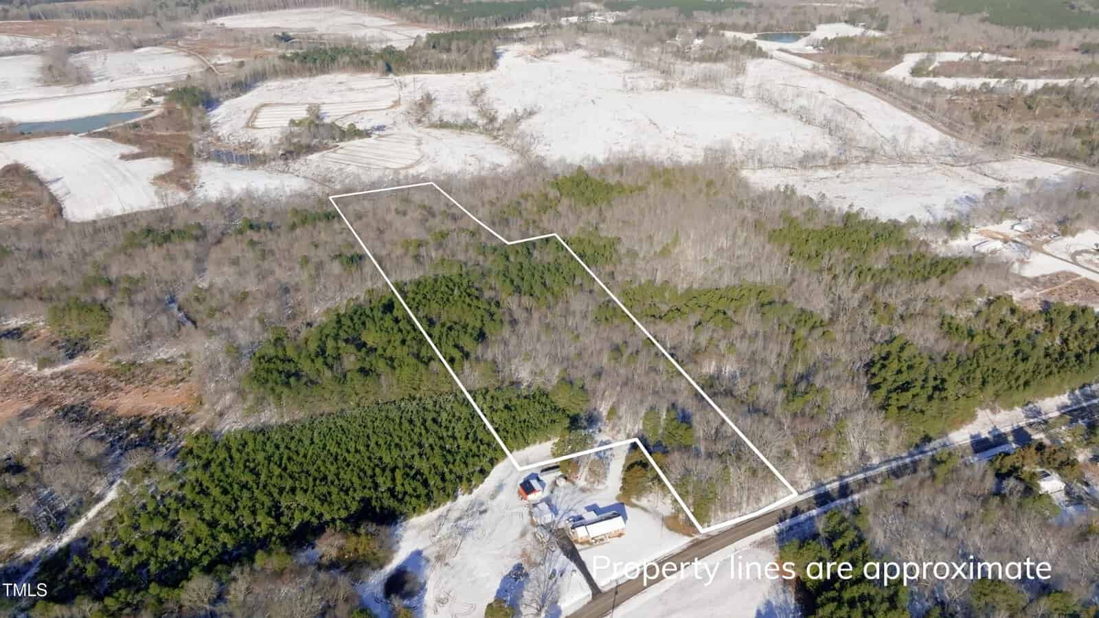 Aerial view of a wooded, rural area showing a parcel of land outlined in white with a few buildings at the bottom; surrounding land has trees and open fields. Text reads: "Property lines are approximate.