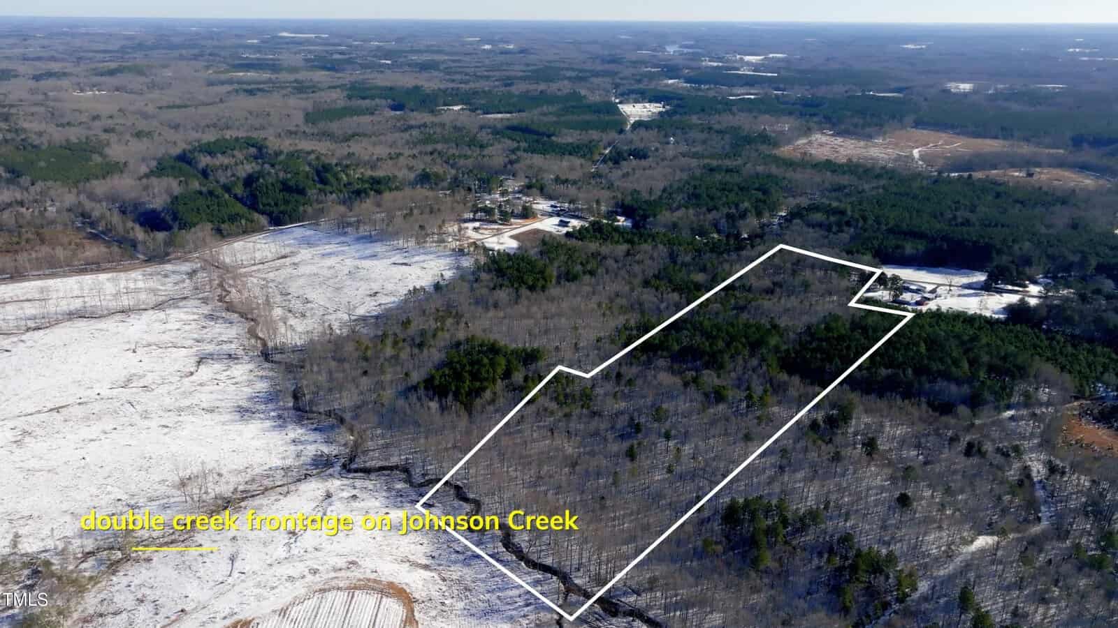 Aerial view of a snowy, wooded landscape with a rectangular plot of land outlined in white, labeled "double creek frontage on Johnson Creek" in yellow text at the bottom left. Some houses and roads are visible in the distance.