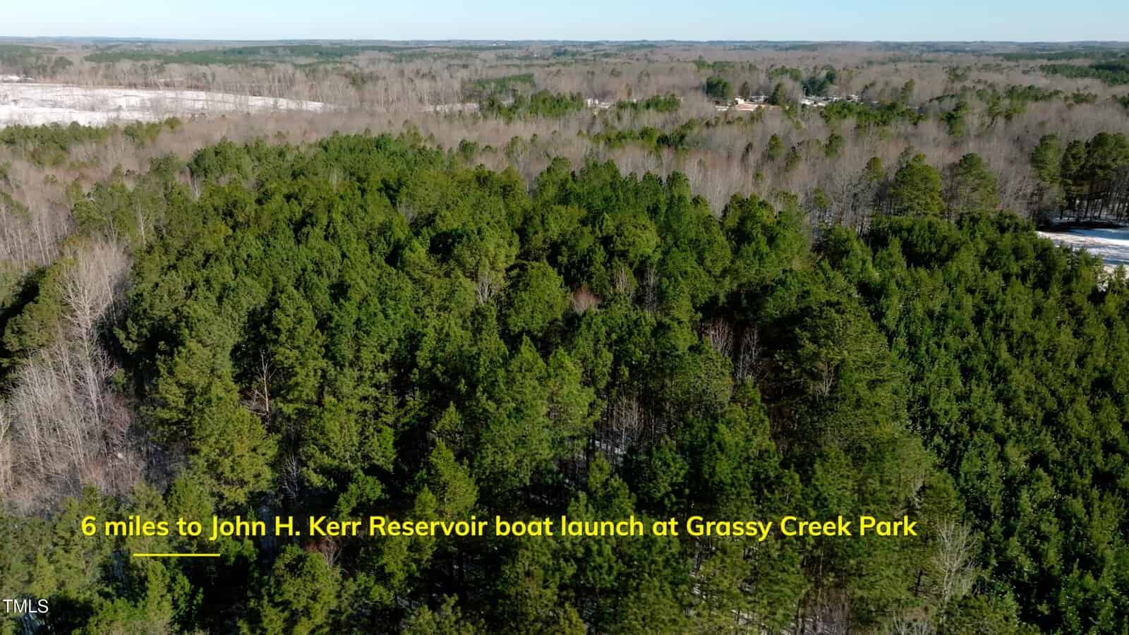 Aerial view of a dense forest with green trees under a clear sky. Yellow text at the bottom reads: "6 miles to John H. Kerr Reservoir boat launch at Grassy Creek Park.