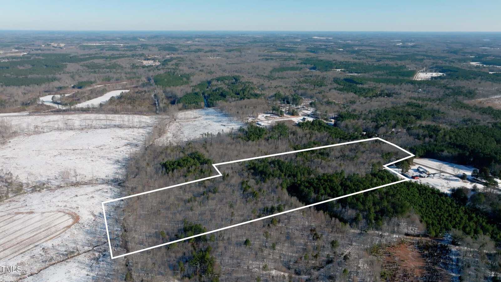 Aerial view of a large, wooded plot of land outlined in white, surrounded by forest and some cleared areas with light snow cover, under a clear sky. Sparse buildings and roads are visible in the distance.