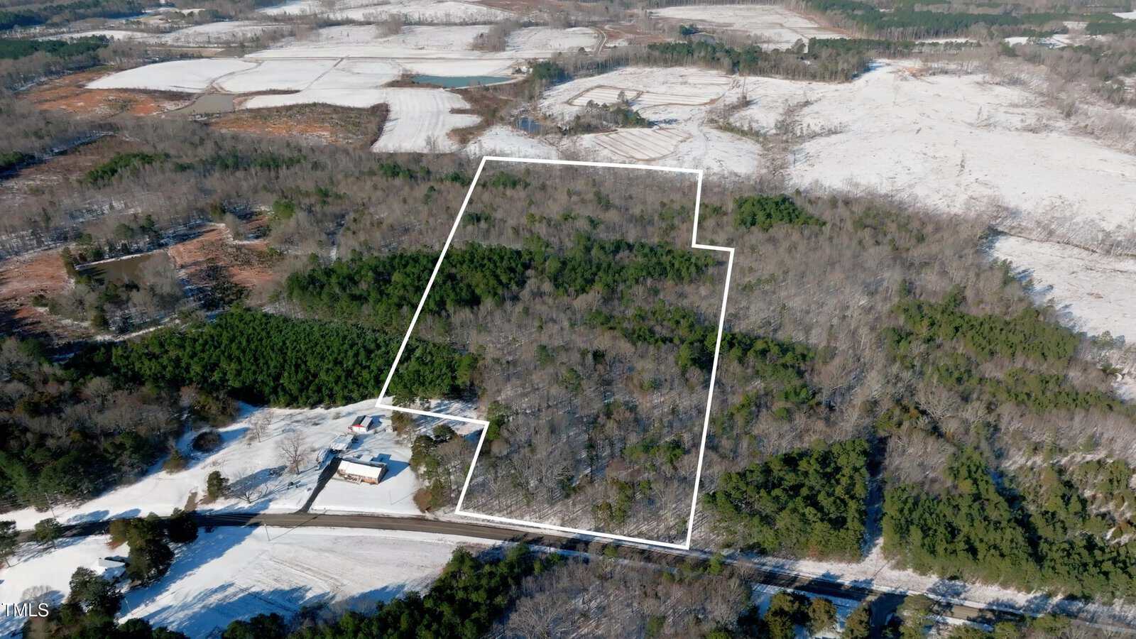 Aerial view of a rural, wooded property outlined in white, surrounded by trees, fields, and patches of snow. A house and driveway are visible near the lower left corner along a road.