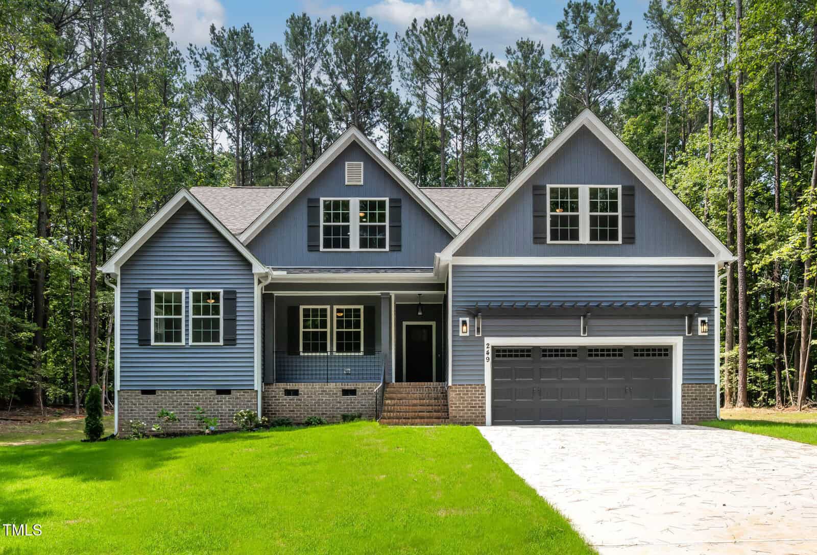 A modern two-story house with blue siding, white trim, and a stone driveway. The home features a covered front porch, double garage, and large windows, surrounded by green lawn and tall trees in the background.