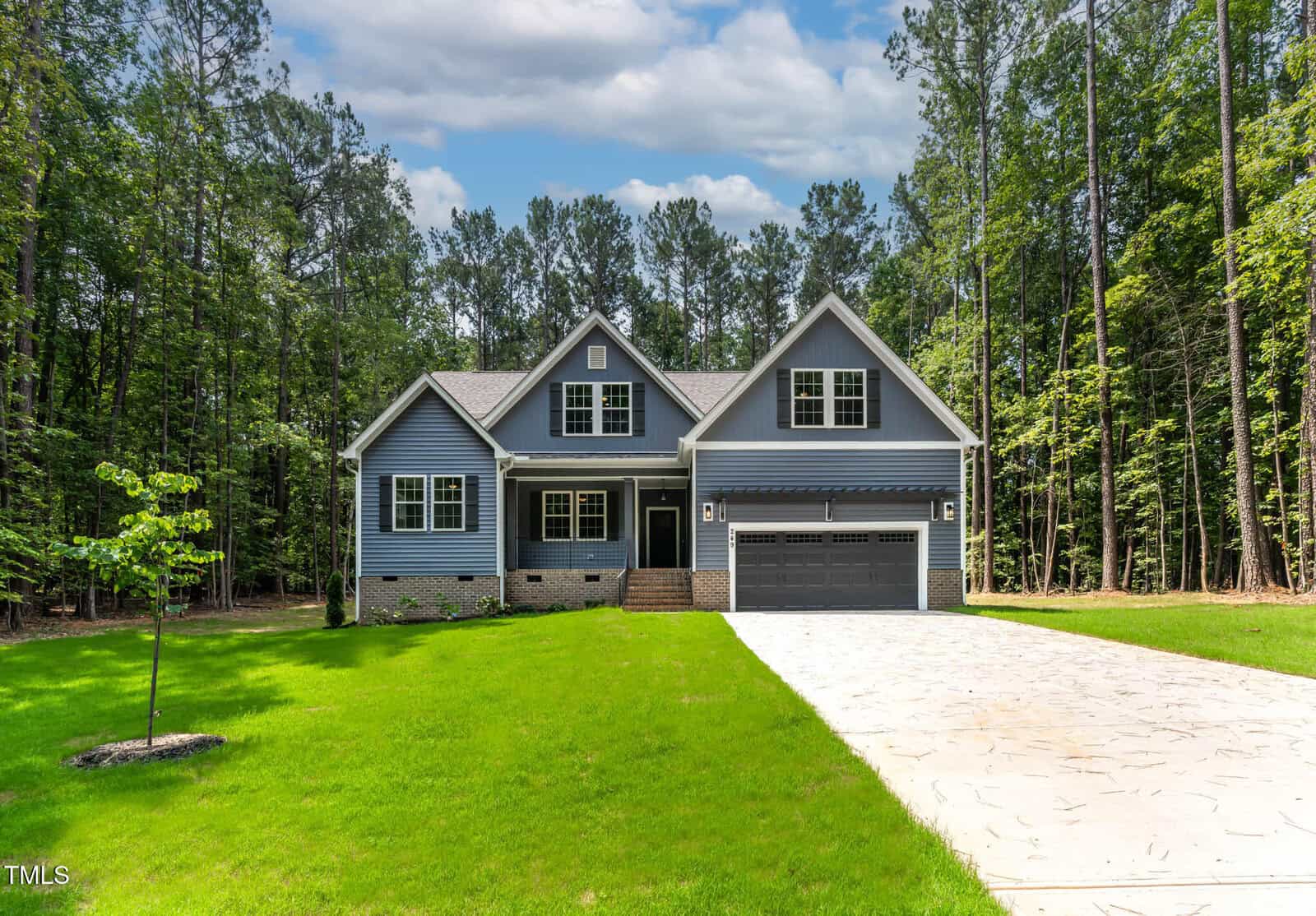 A modern two-story house with blue siding, a large driveway, and a two-car garage sits surrounded by lush green grass and tall trees under a partly cloudy sky.