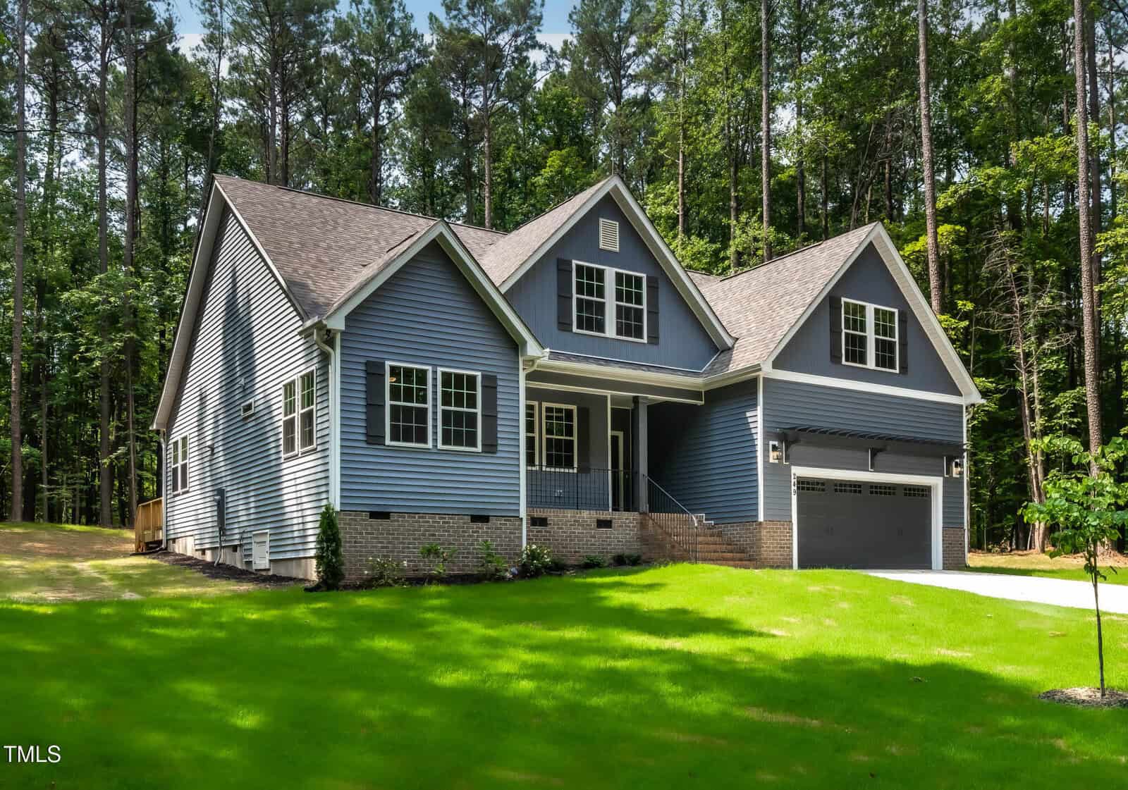 A blue two-story suburban house with white trim, a covered front porch, and a two-car garage, set on a large green lawn surrounded by tall trees.