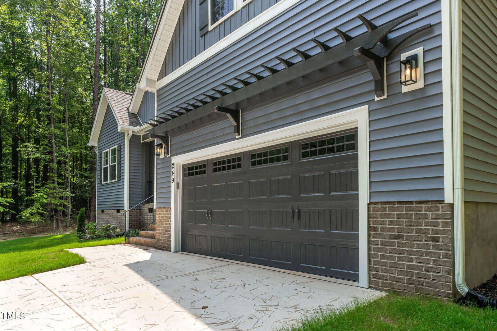 A modern house with blue siding, brick accents, a two-car garage with dark doors, black pergola trim, and outdoor lantern lights. The driveway is concrete and surrounded by green trees and a lawn.