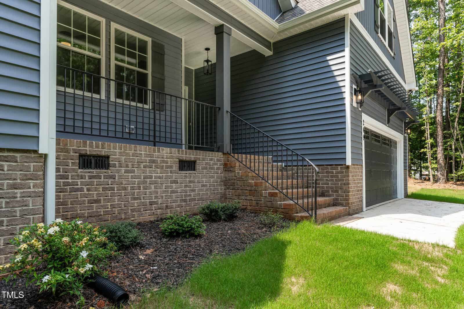A view of a modern house exterior with blue siding, brick foundation, a black railing on the porch steps, double garage doors, and landscaped yard with grass and small shrubs.