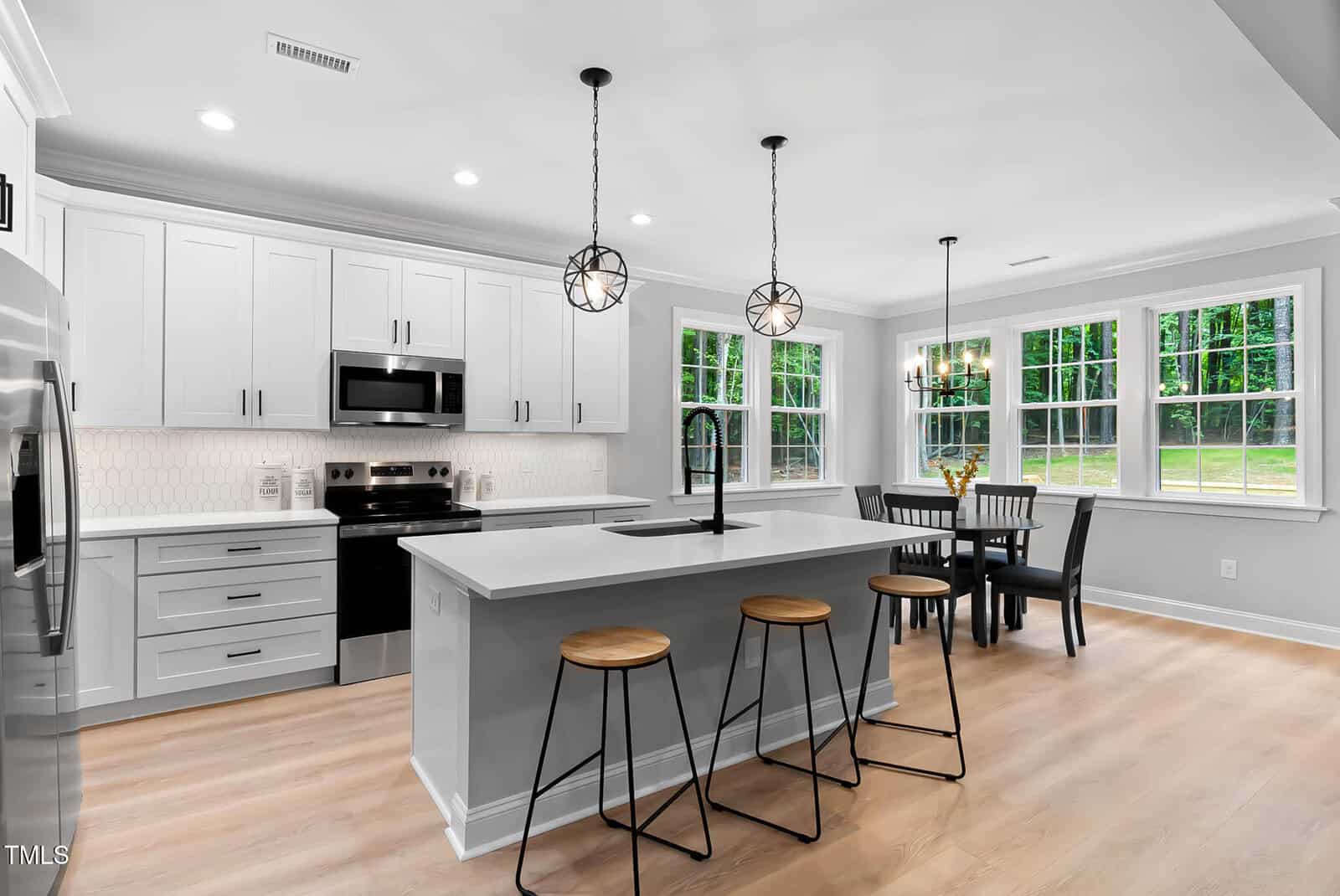Modern kitchen with white cabinets, stainless steel appliances, a large island with three wooden stools, pendant lights above the island, and a dining area with a table and chairs near large windows.