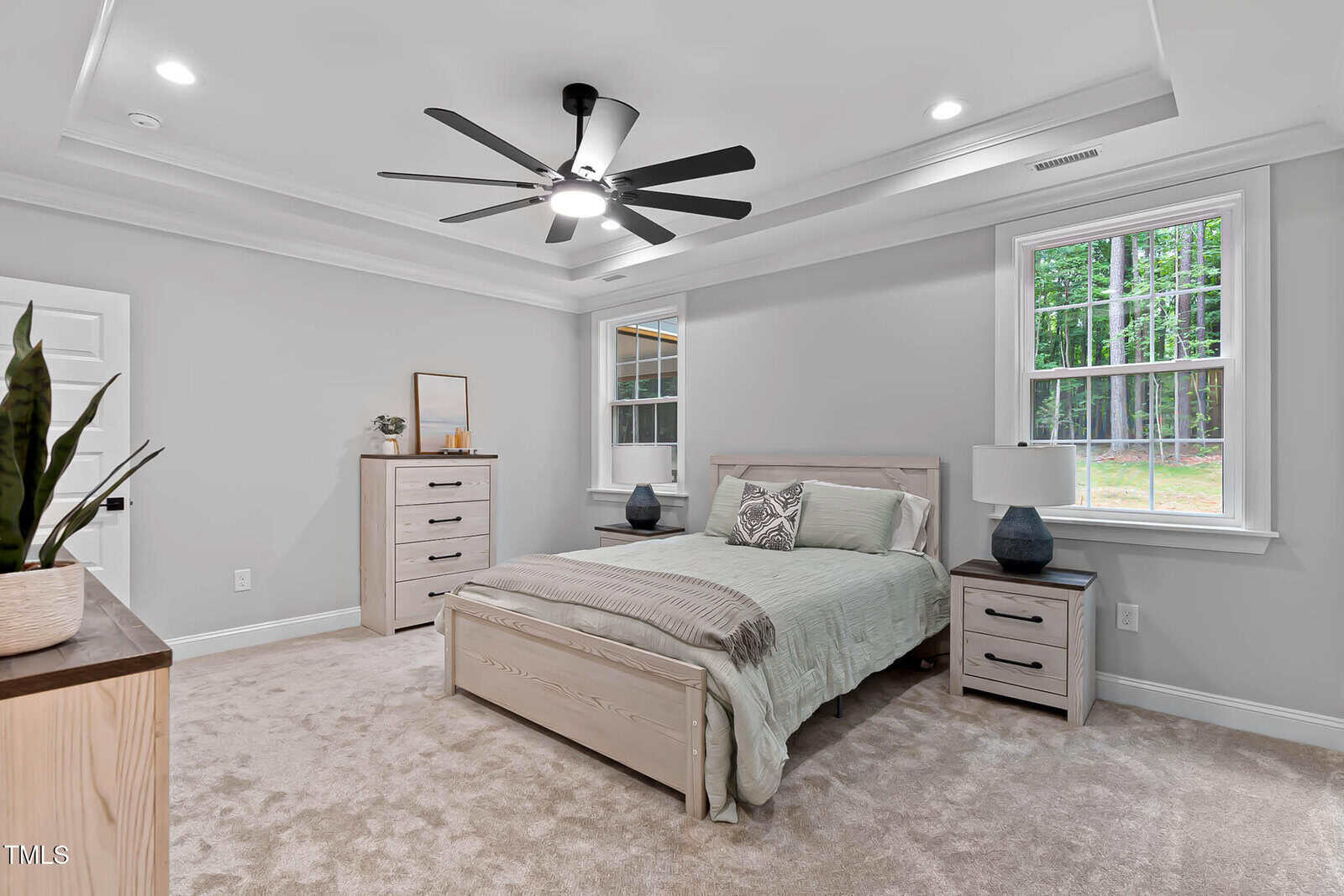 Bedroom with light wood furniture, neutral bedding, two windows with outdoor views, carpeted floor, ceiling fan with light, and minimal decor including a plant, lamp, and mirror. Walls are light gray.