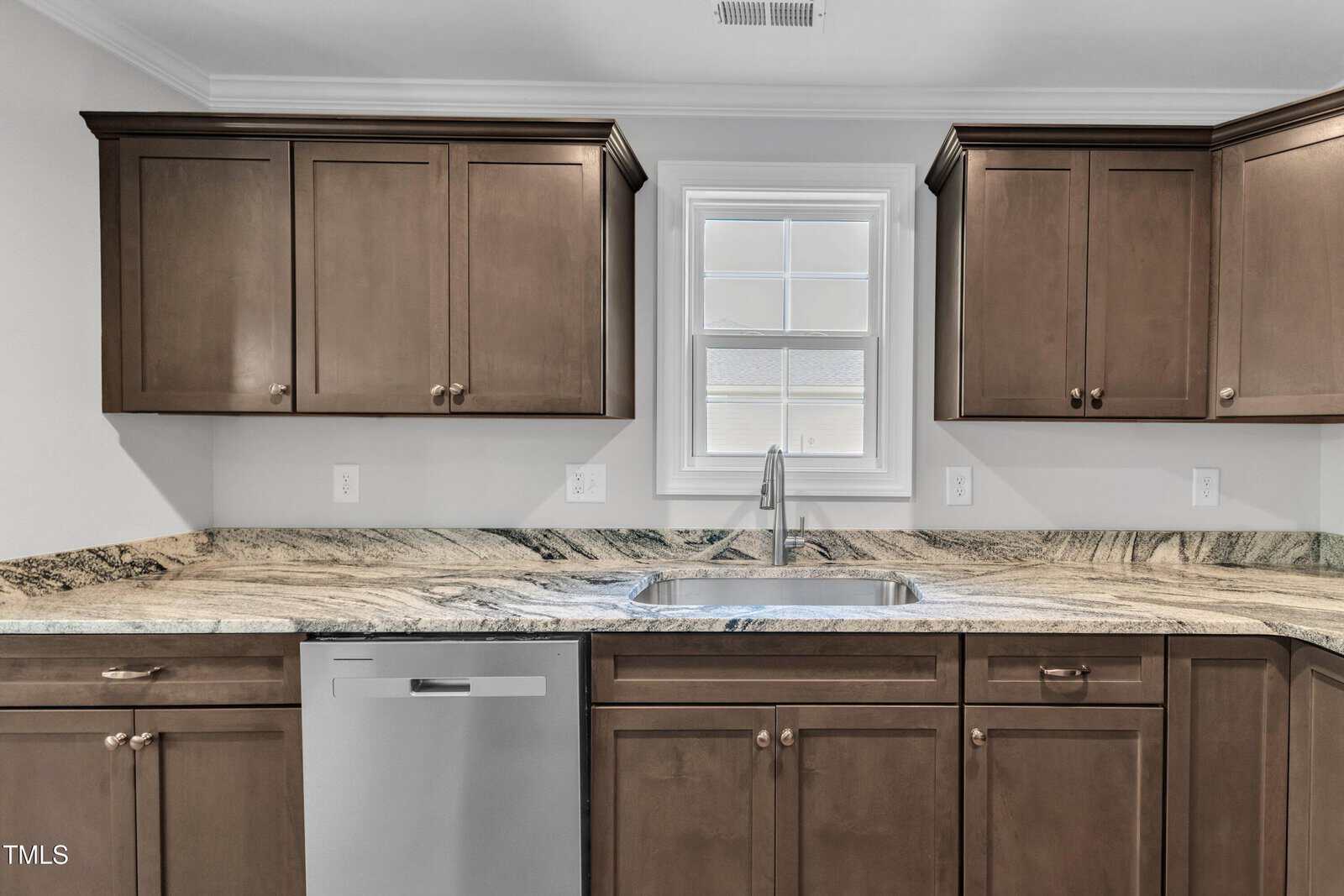 Kitchen with dark wood cabinets, granite countertop, stainless steel sink, and dishwasher beneath a window. White walls, electrical outlets, and vent on ceiling are also visible.