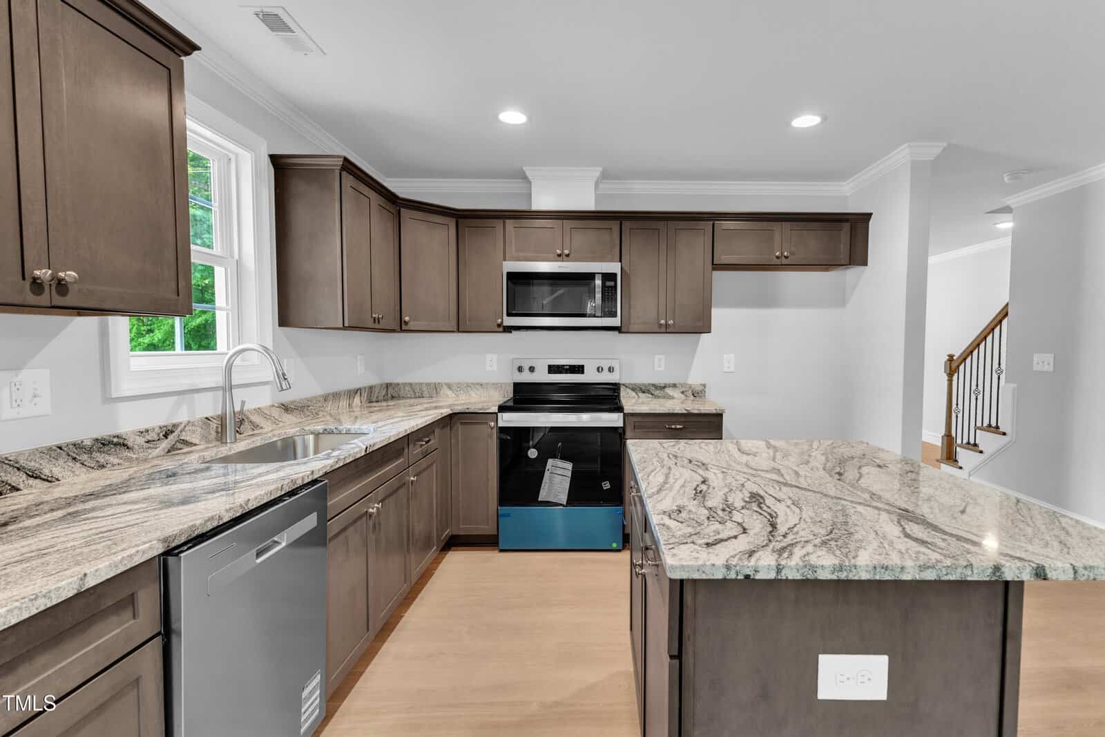 Modern kitchen with dark wood cabinets, stainless steel appliances, marble countertops, a center island, a large window above the sink, light wood flooring, and an open layout leading to a staircase.