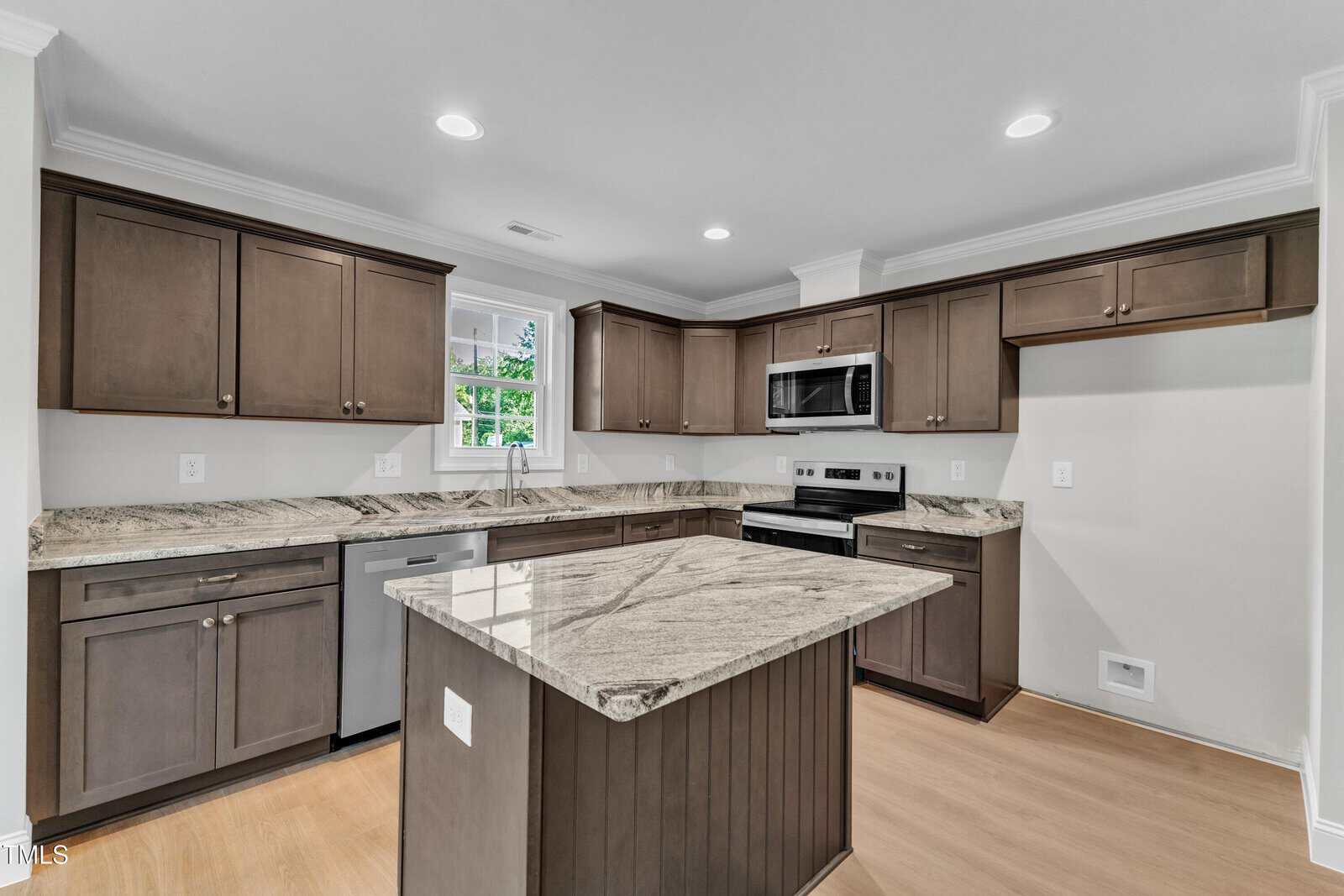 Modern kitchen with dark wood cabinets, stainless steel appliances, marble countertops, center island, light wood flooring, and a window above the sink, all under recessed ceiling lights.