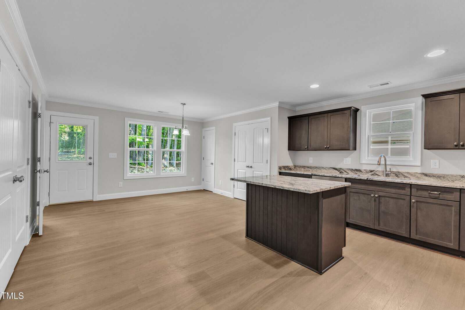 Empty kitchen and dining area with wood floors, dark cabinets, granite countertops, island, and multiple windows letting in natural light; white walls and doors, with recessed and pendant lighting visible.