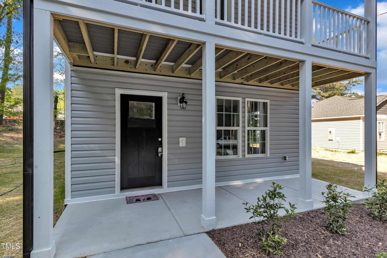 A modern gray house exterior with a black front door, white trim, and a covered front porch supported by white columns. There is a window beside the door and small shrubs planted along the walkway.
