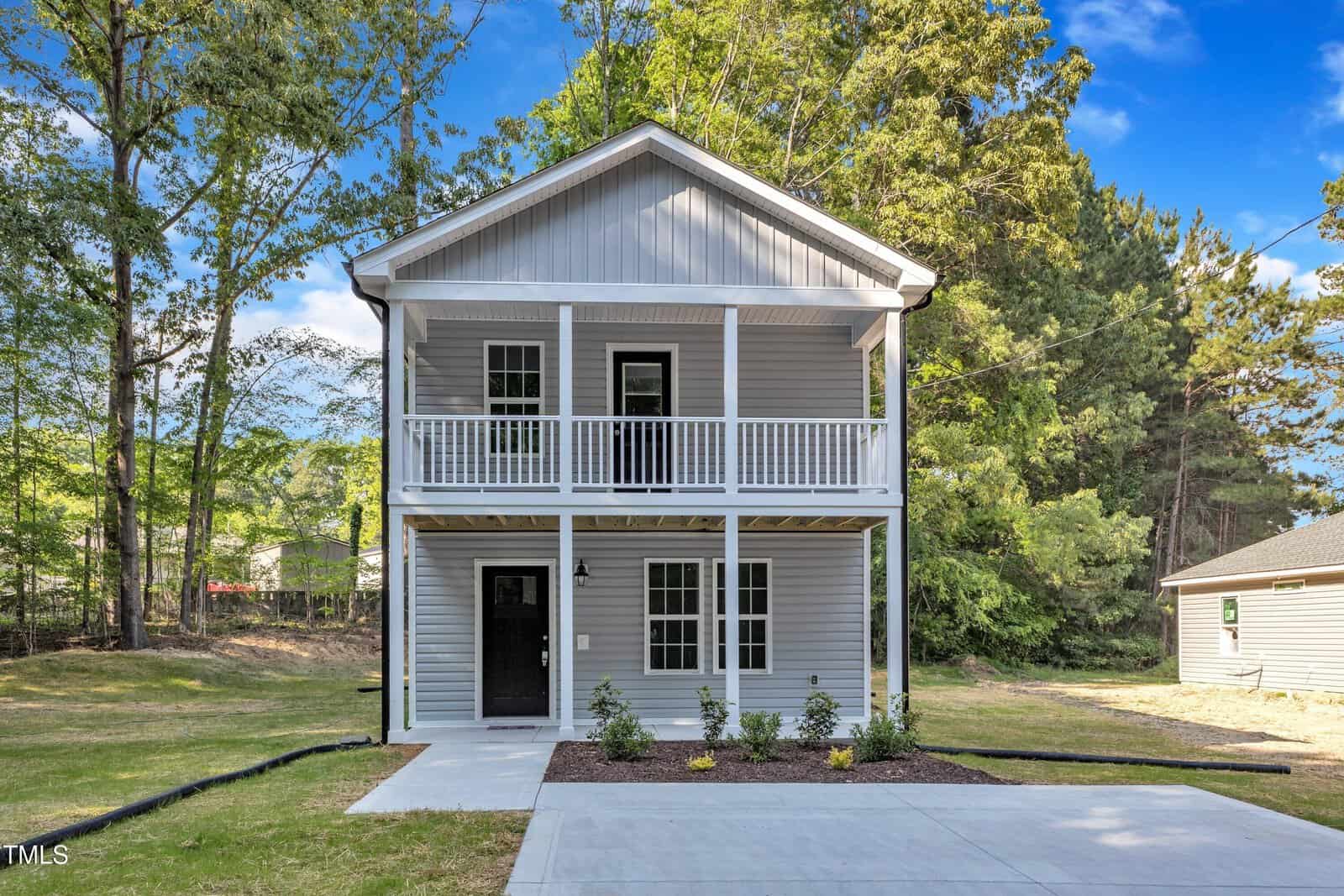 Two-story gray house with white trim, covered upper and lower porches, black doors, small shrubs in front, concrete driveway, and surrounded by green trees under a blue sky.