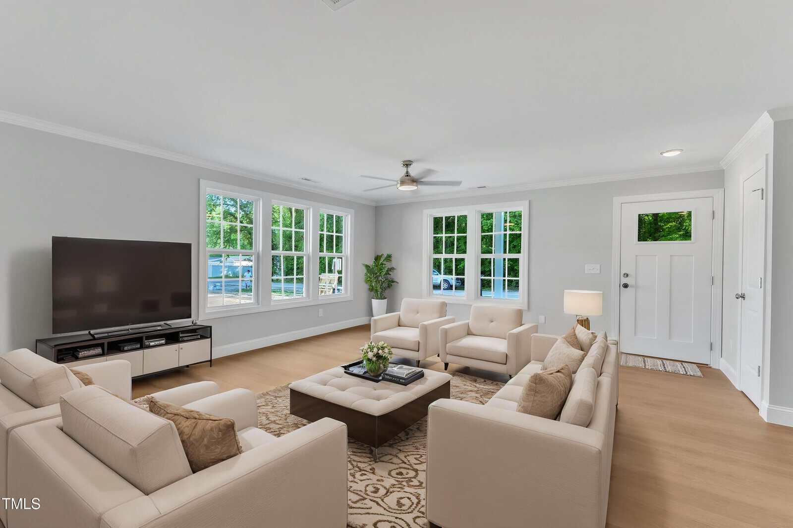 A bright living room with beige sofas arranged around a square ottoman, a large TV on a stand, light wood flooring, white walls, multiple windows, and a white front door.
