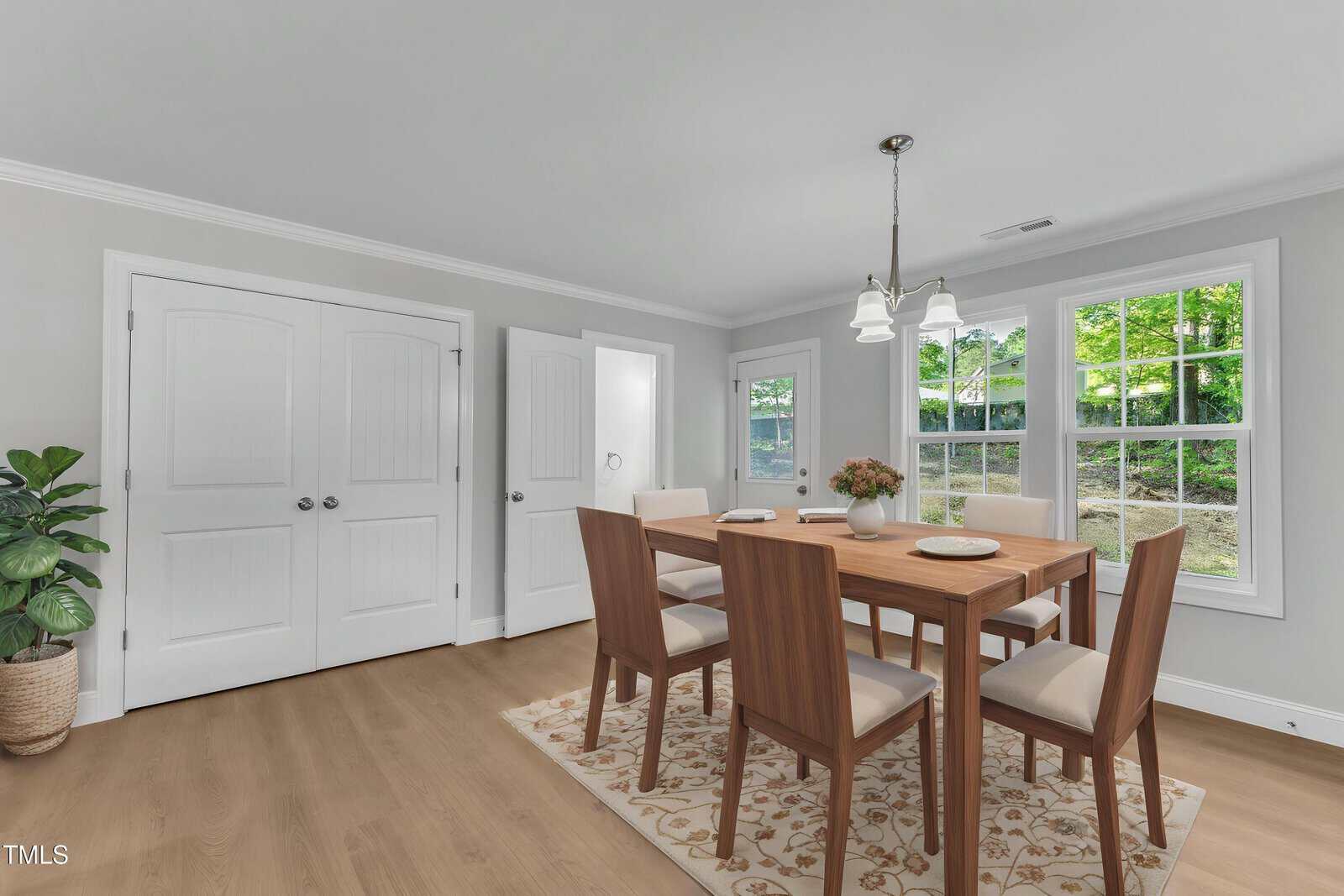 A bright dining room with light gray walls, wood flooring, a wooden table with four chairs, a floral rug, a potted plant, and large windows showing greenery outside. A double-door closet is on the left.