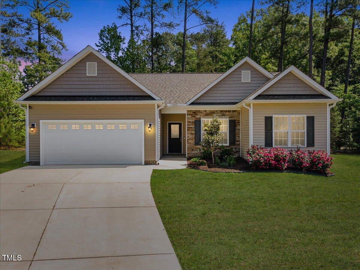 Charming single-story house at 217 Griffis Drive with a double garage, gray siding, stone accents, front door with window, manicured lawn, blooming pink bushes, and tall trees stemming up to a clear blue sky.