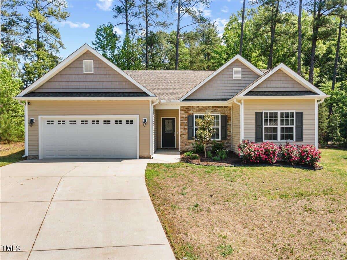 Single-story suburban house with a two-car garage, gray siding, and stone accents. There are pink flowering bushes in front, a concrete driveway, and trees in the background under a blue sky.