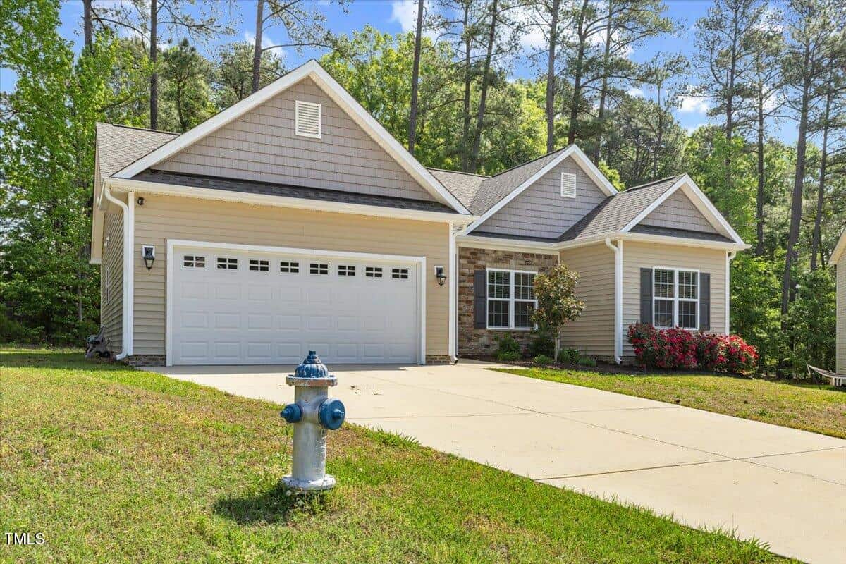 Single-story beige house with a two-car garage, gabled roof, and small front yard. A blue fire hydrant is on the grass near the driveway. There are red bushes by the front window and trees in the background.