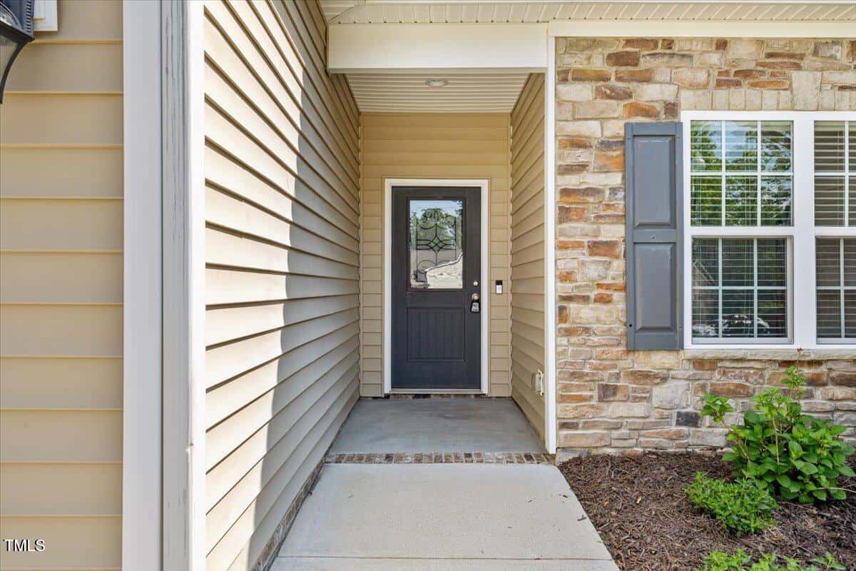 A house entryway with beige horizontal siding on the left, tan stonework on the right, a black front door with glass panes, white trim, and a small concrete porch with a garden bed along the side.