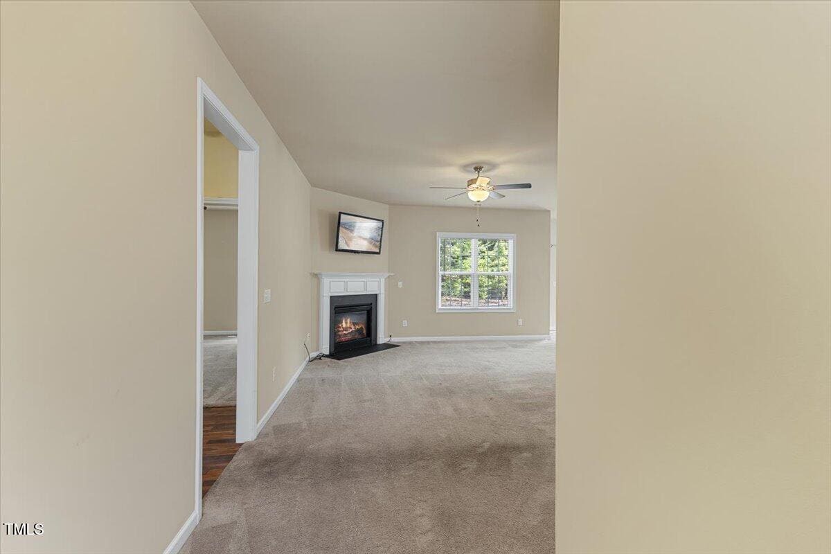 Carpeted living room with beige walls, a ceiling fan with light, a window, a fireplace with a TV above it, and entrances to adjacent rooms. The space is empty and well-lit with natural light.