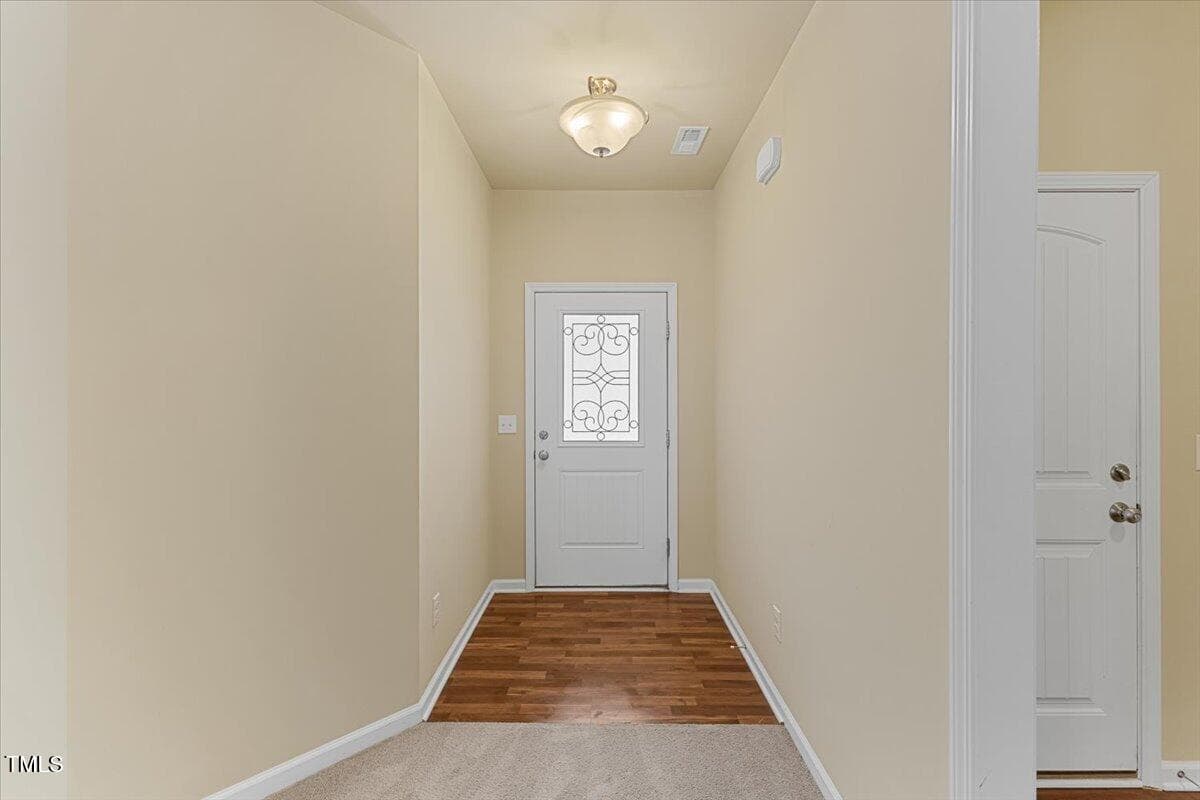 A hallway with beige walls and a ceiling light leads to a white door with a decorative glass panel. The floor transitions from carpet to wood near the door. A second white door with silver handles is partially visible on the right.