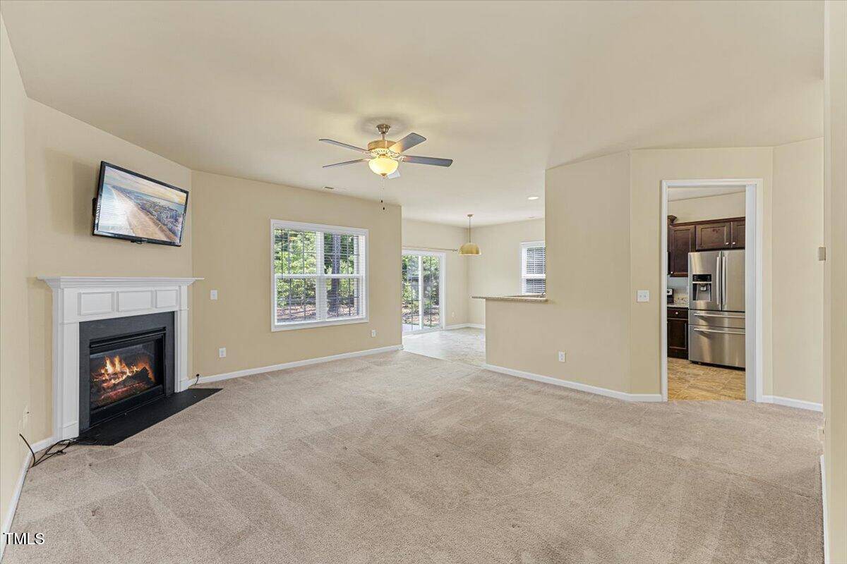 A spacious living room with beige walls and carpet, a ceiling fan, a fireplace with a TV above it, and large windows. The room opens to a tiled dining area and kitchen with stainless steel appliances.