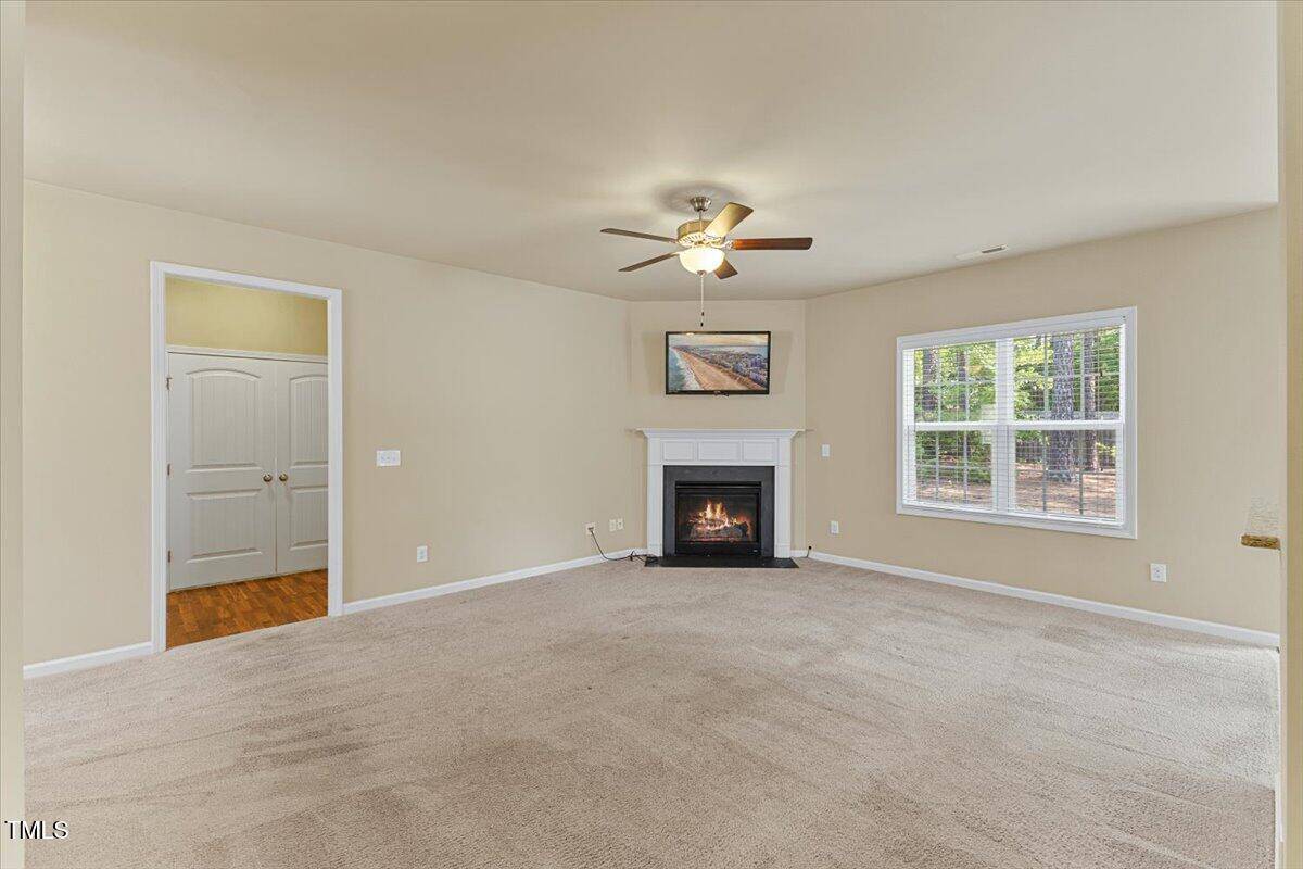 A spacious living room with beige carpet, light-colored walls, a ceiling fan with a light, a white fireplace with a painting above it, and a large window letting in natural light. Double doors lead to another room.