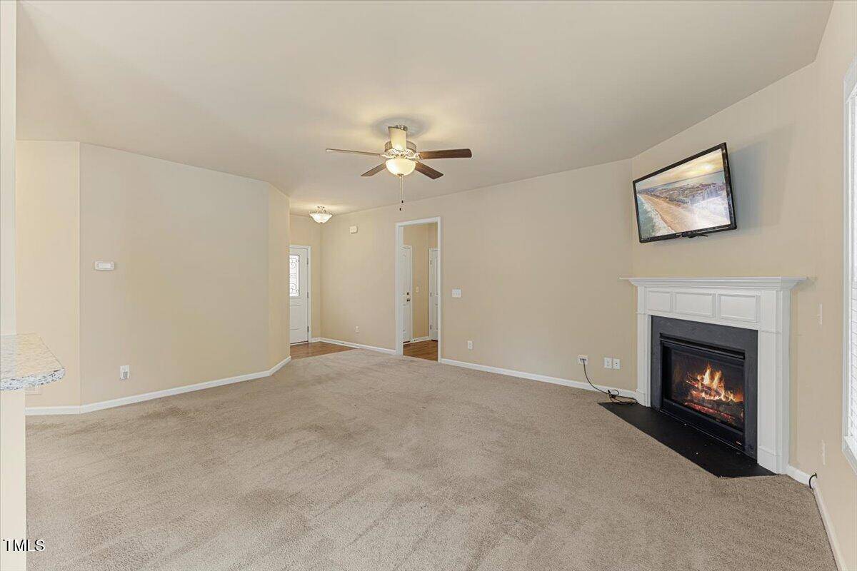 A spacious living room with beige walls and carpet, a ceiling fan, a corner fireplace with a lit fire, and a flat-screen TV mounted above the mantel. There is a hallway and a doorway visible in the background.