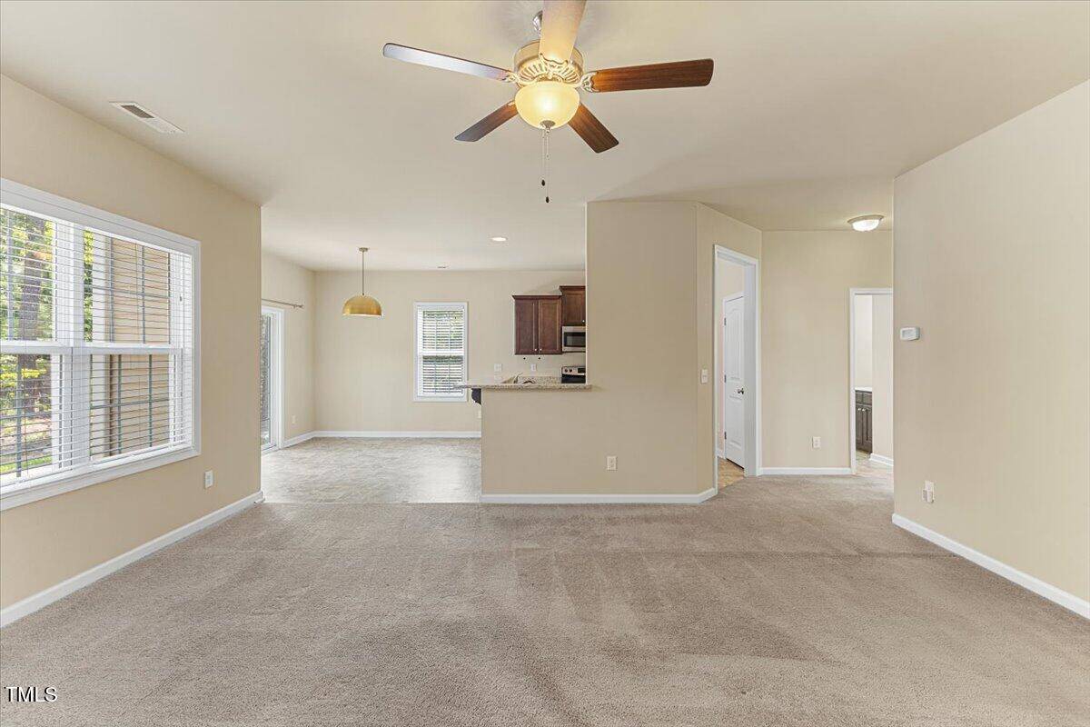 A spacious living room with beige carpet, ceiling fan, and neutral walls, opening to a tiled dining area and kitchen with brown cabinets, stainless steel appliances, and several large windows for natural light.