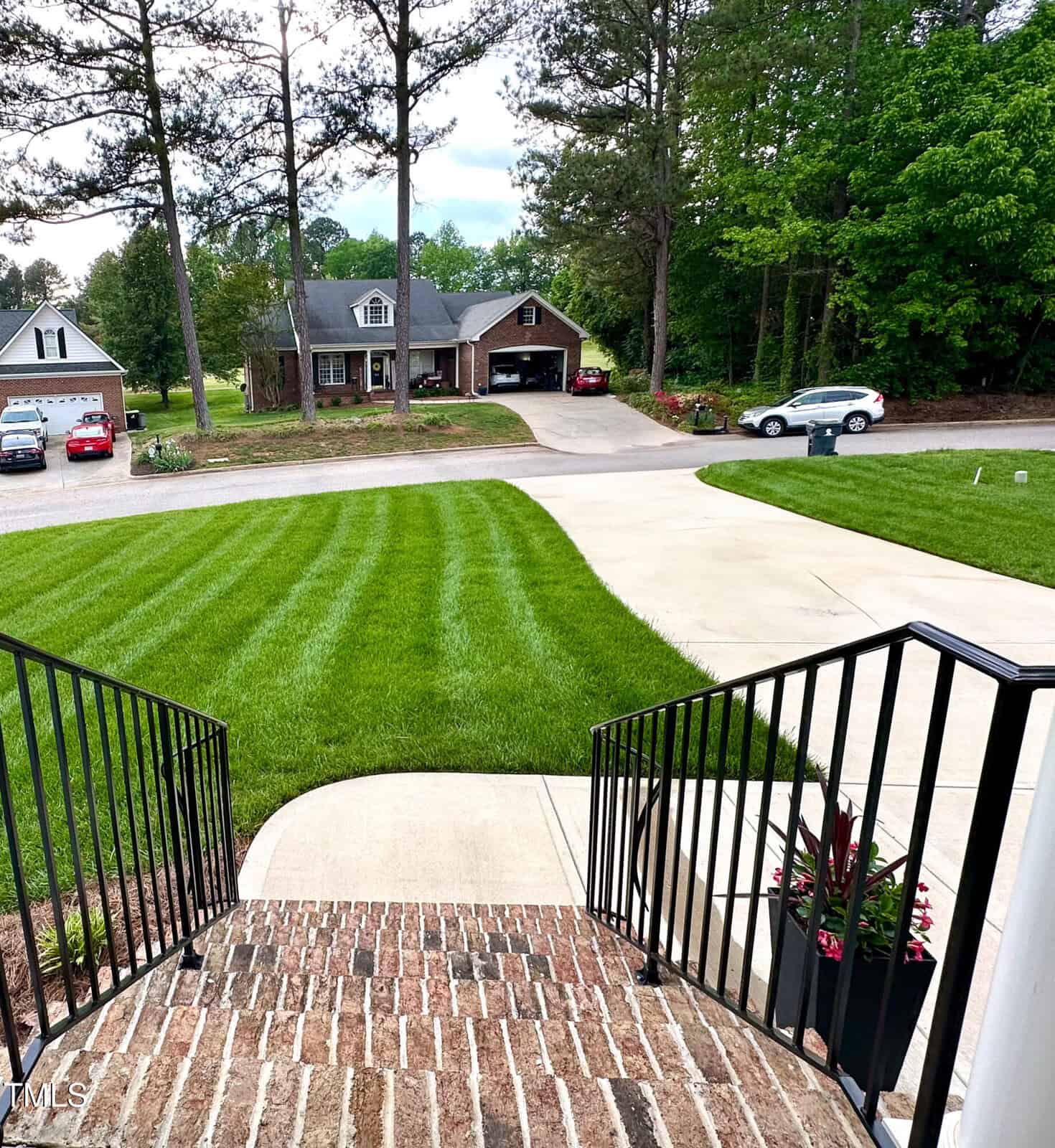 A view from brick steps with black railings, overlooking a neatly mowed lawn with striped patterns, a curved concrete driveway, cars parked, and houses surrounded by trees.