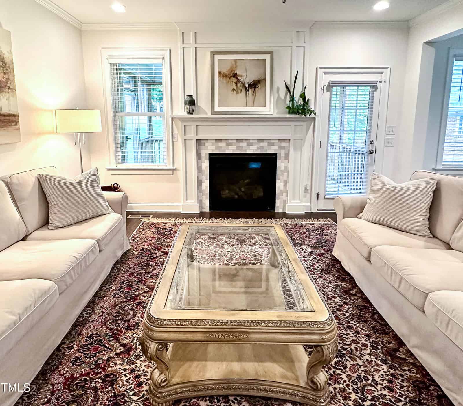 A living room with two cream sofas facing each other, a glass-top ornate coffee table, a fireplace with a white mantel, plants, artwork above the mantel, and a patterned rug on the floor.