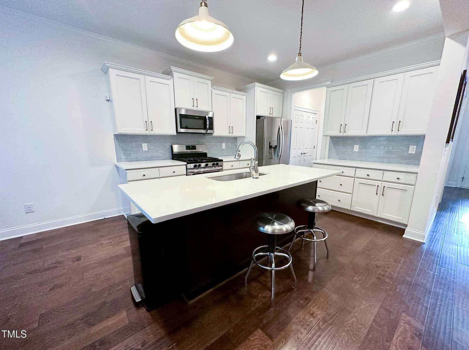 Modern kitchen with white cabinets, stainless steel appliances, a large island with a sink, and two metal stools. Pendant lights hang above the island, and the floor is dark wood.