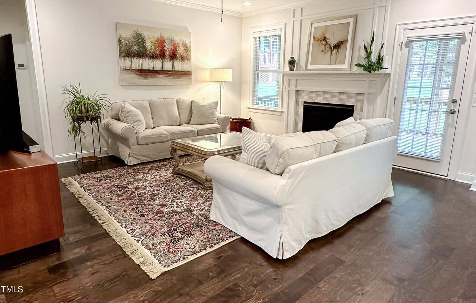 A living room with two white sofas, a glass coffee table, a patterned rug, and a fireplace. There is artwork above the fireplace and sofa, plants, a floor lamp, and windows letting in natural light.