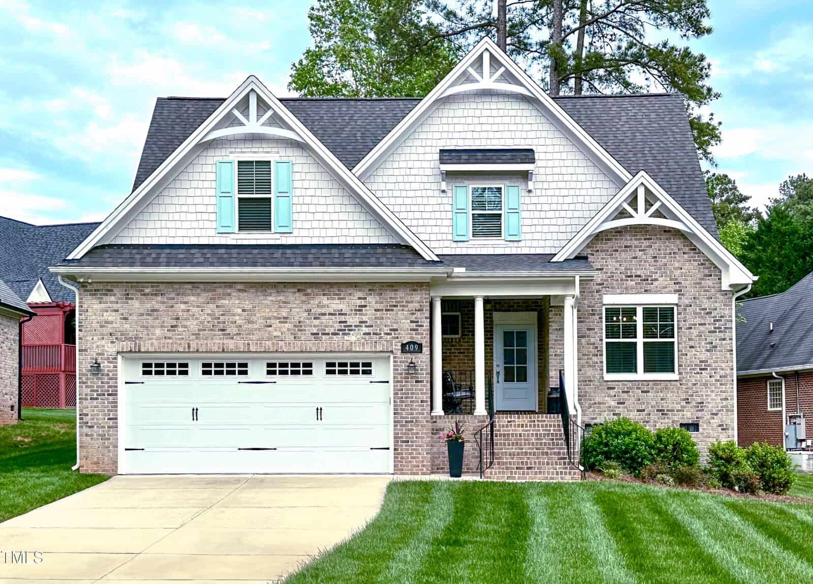 A two-story suburban house at 409 Eagle Court with a light brick exterior, white trim, gray shingles, teal window shutters, a covered front porch, and double garage. The green lawn blends into the inviting Henderson neighborhood.