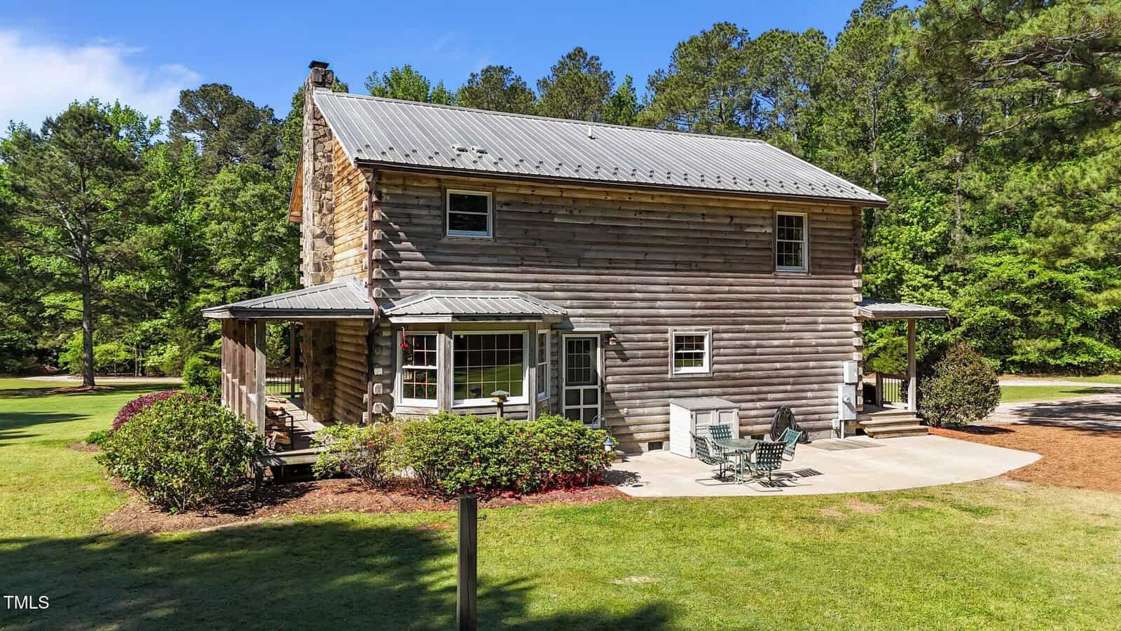 A rustic two-story log cabin with a metal roof sits on a well-manicured lawn, surrounded by trees. There is a patio with outdoor furniture and a wraparound porch with bushes and shrubs nearby.