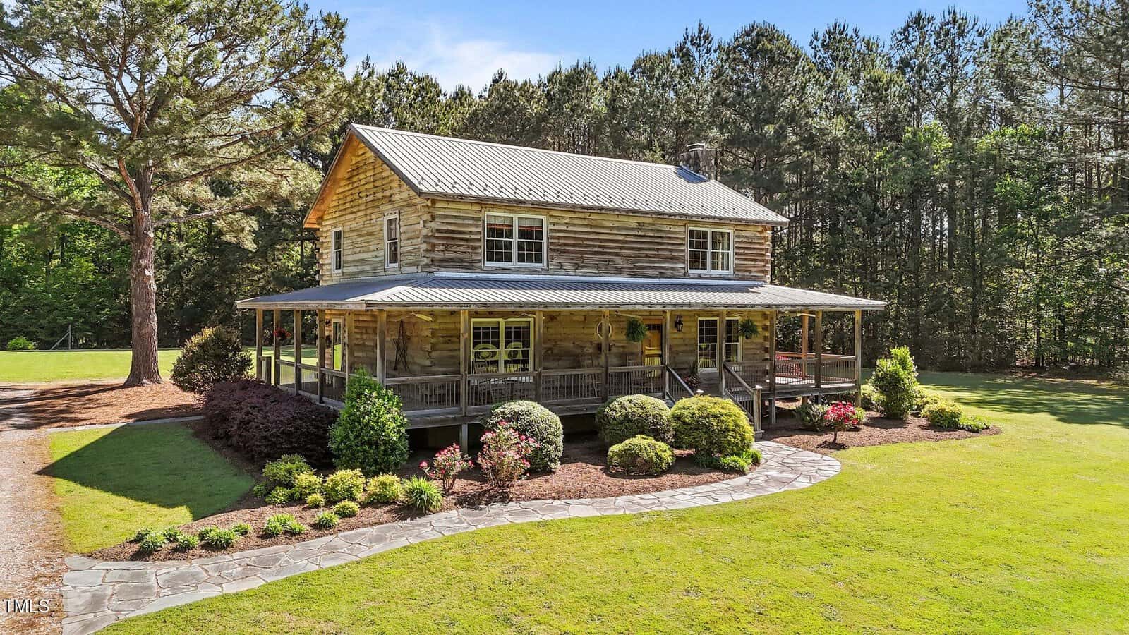 A two-story log cabin with a wraparound porch sits on a well-manicured lawn, surrounded by trees and landscaped bushes, under a bright blue sky.