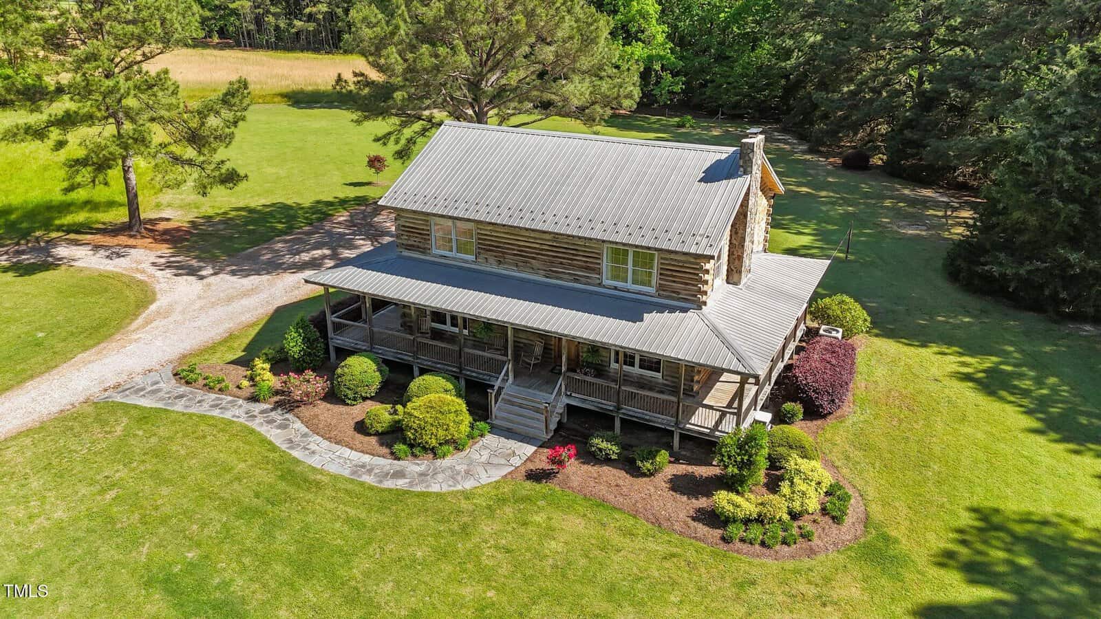 Aerial view of a rustic log cabin with a metal roof and wraparound porch, surrounded by manicured lawn, shrubs, and trees, with a stone walkway leading to the porch.