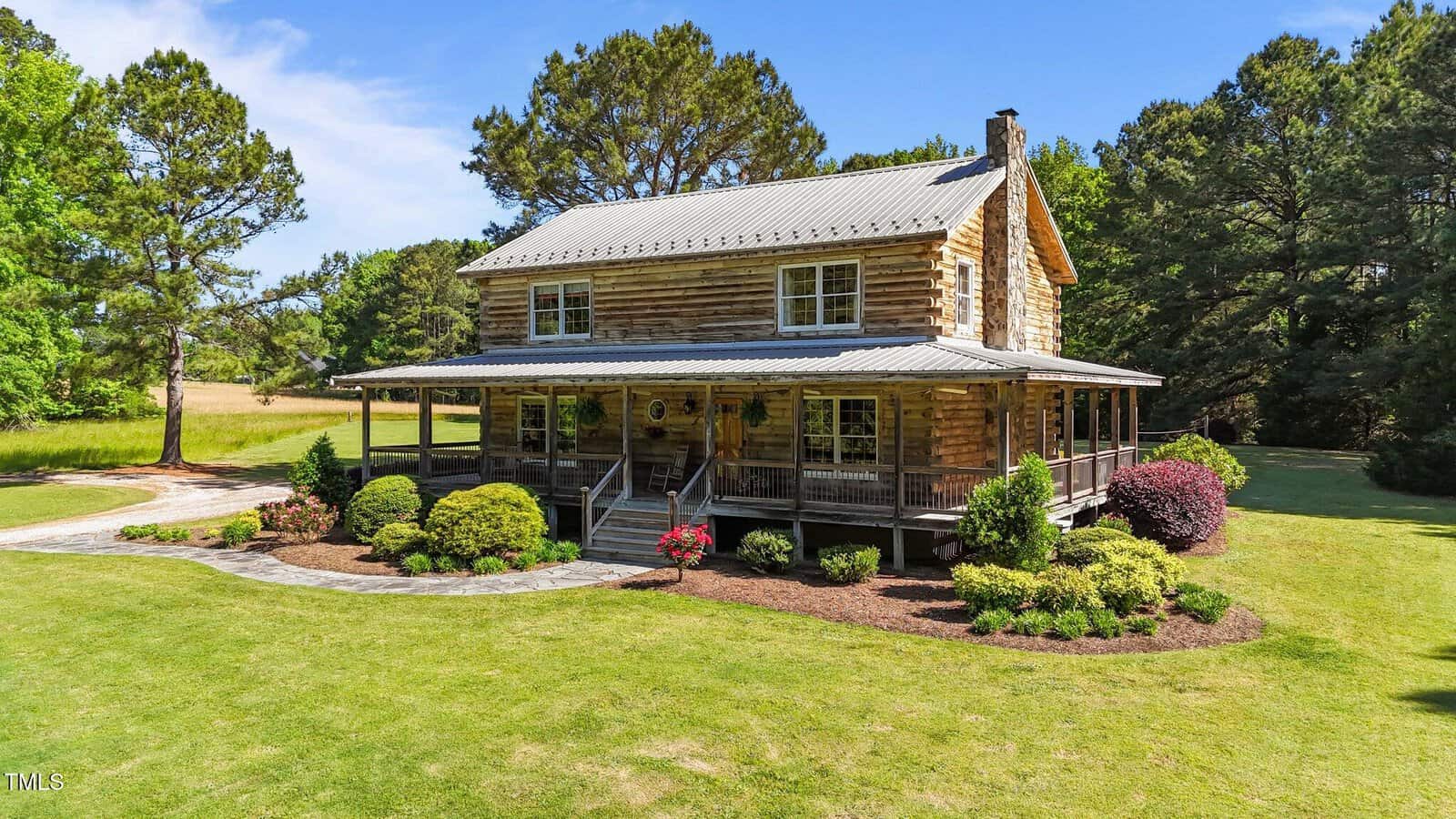A two-story log cabin with a metal roof and wraparound porch sits on a well-maintained lawn, surrounded by trees, shrubs, and landscaping on a sunny day.