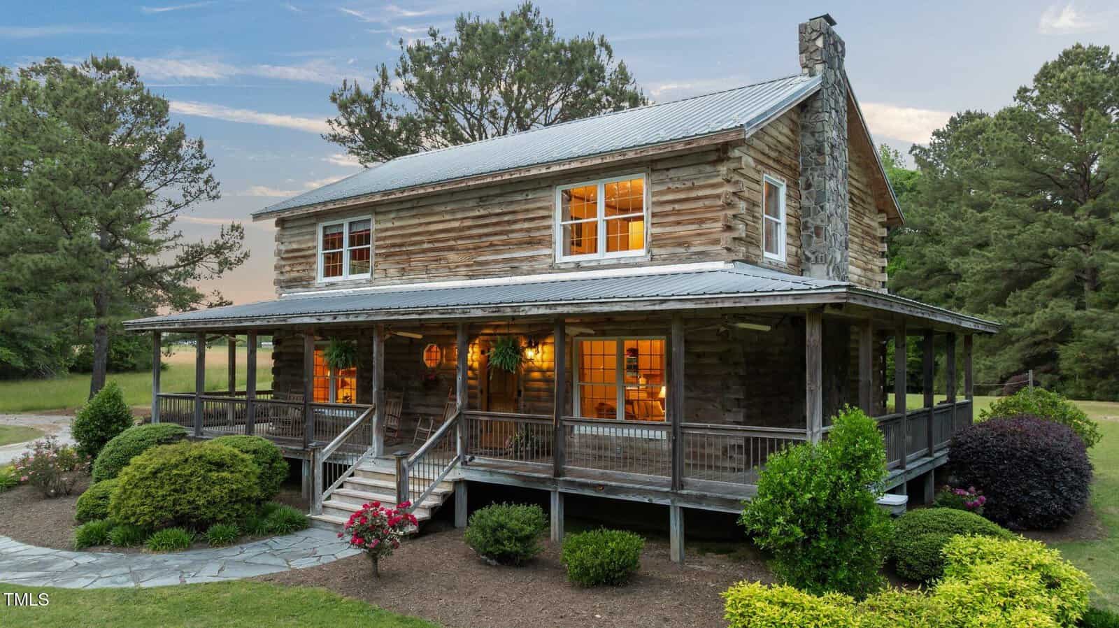 A rustic two-story log cabin with a metal roof, large stone chimney, and a wraparound porch is surrounded by bushes, flowers, and trees under a partly cloudy sky at dusk. Warm lights glow from the windows.