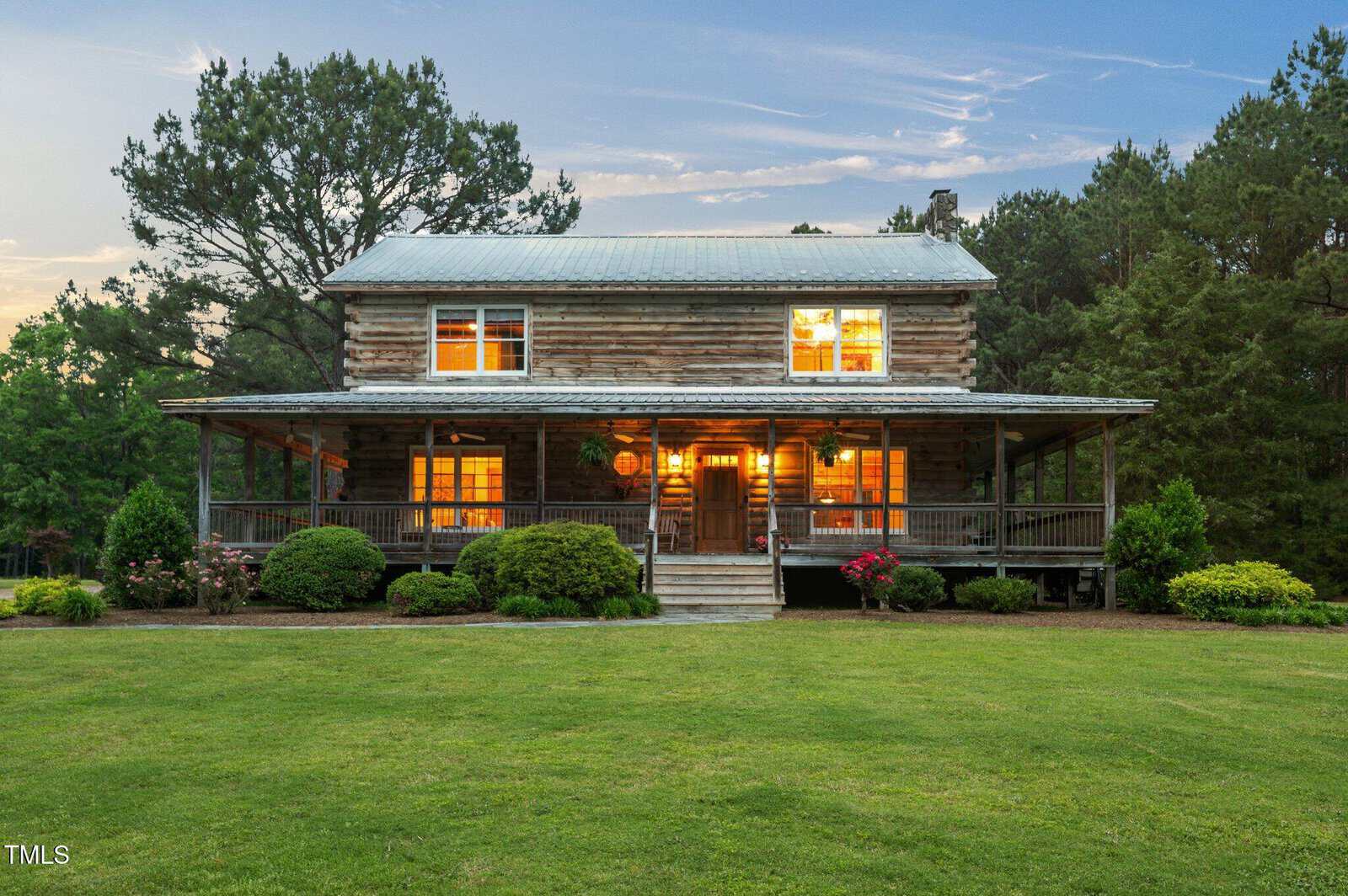 A two-story log cabin with a metal roof and wraparound porch, surrounded by green lawn, shrubs, and trees. Warm lights shine from the windows at dusk.