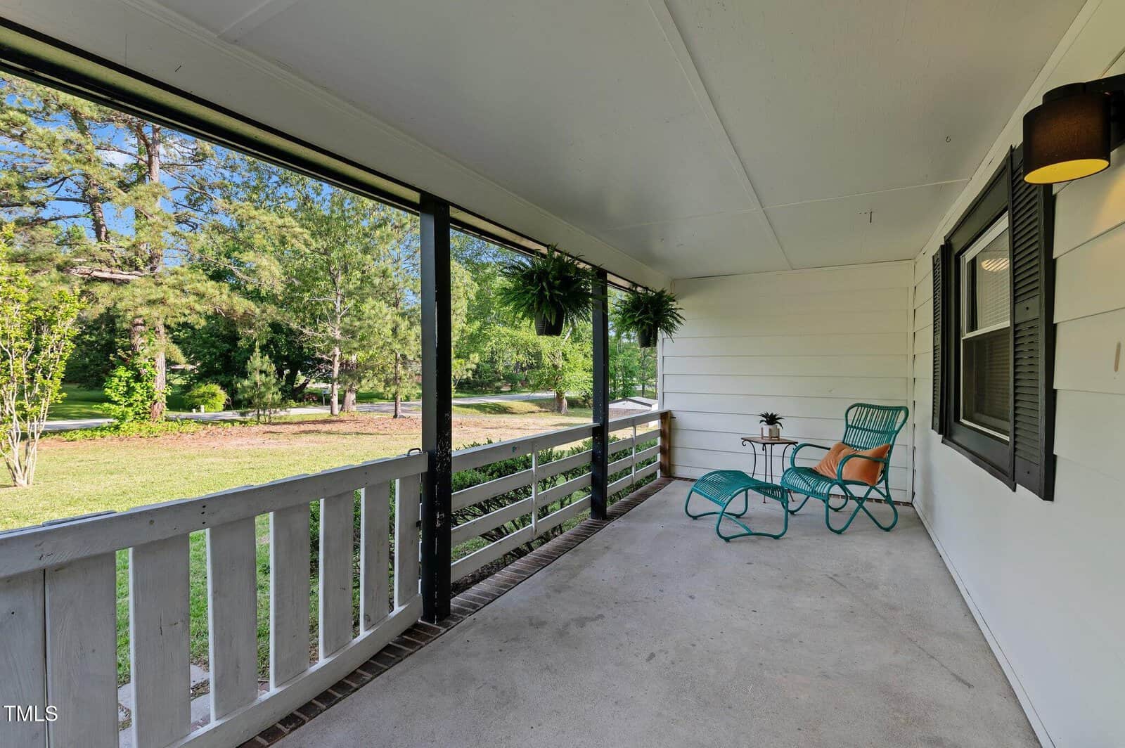 A spacious covered front porch with white railing, two teal chairs, a small table with a plant, hanging ferns, and a view of a green lawn with trees in the background.