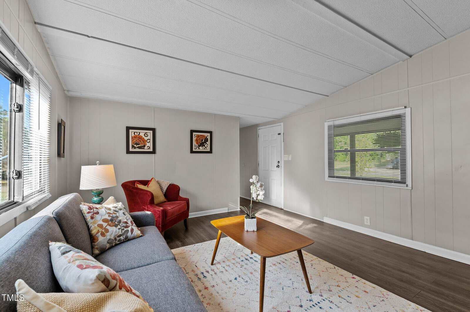 A living room with a gray sofa, red armchair, wooden coffee table, area rug, and artwork on light-colored walls. There is a window with blinds and another next to the white entry door. The flooring is dark wood.